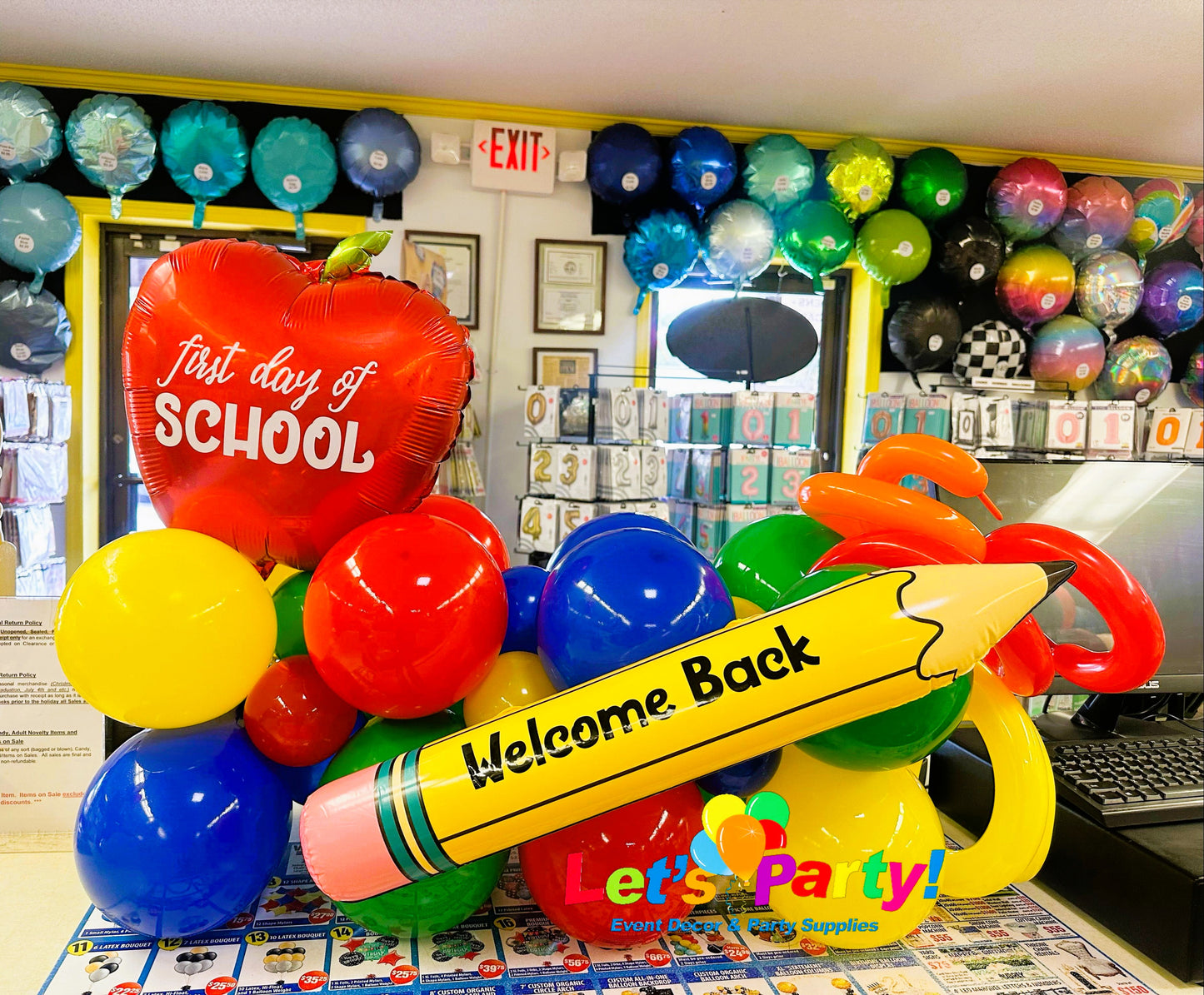 A colorful balloon decoration set shaped like school supplies, including a large yellow pencil with 'Welcome Back' written on it, a red apple balloon reading 'first day of SCHOOL,' and several red, blue, yellow, green, and orange balloons in Let's Party! store.