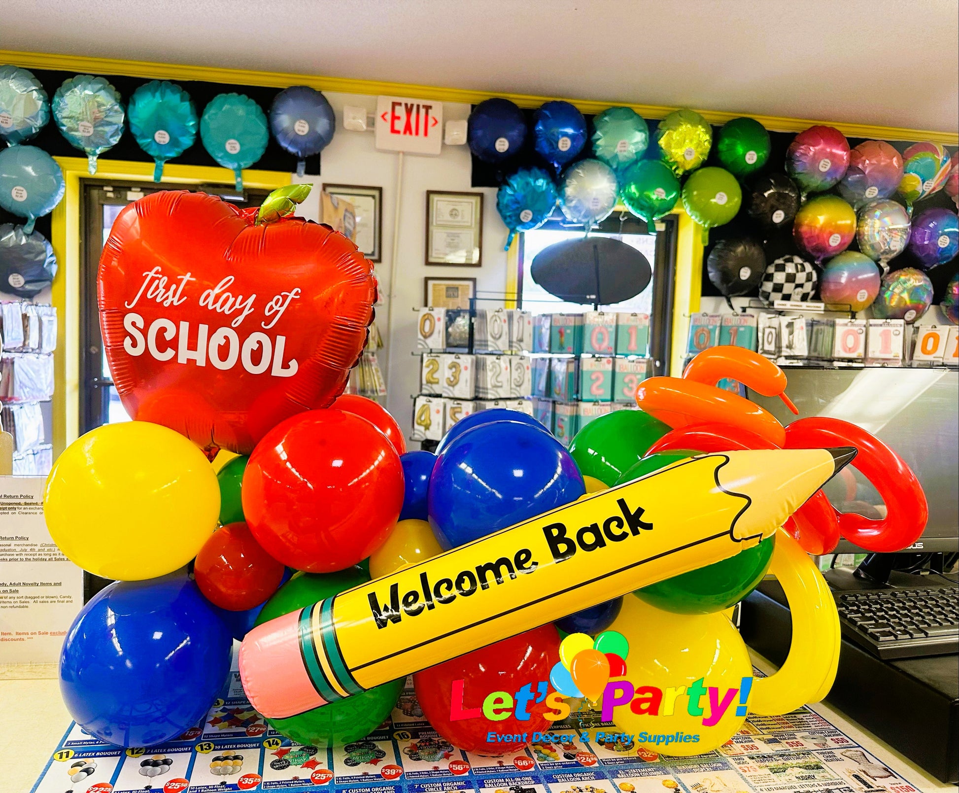 A colorful balloon decoration set shaped like school supplies, including a large yellow pencil with 'Welcome Back' written on it, a red apple balloon reading 'first day of SCHOOL,' and several red, blue, yellow, green, and orange balloons in Let's Party! store.
