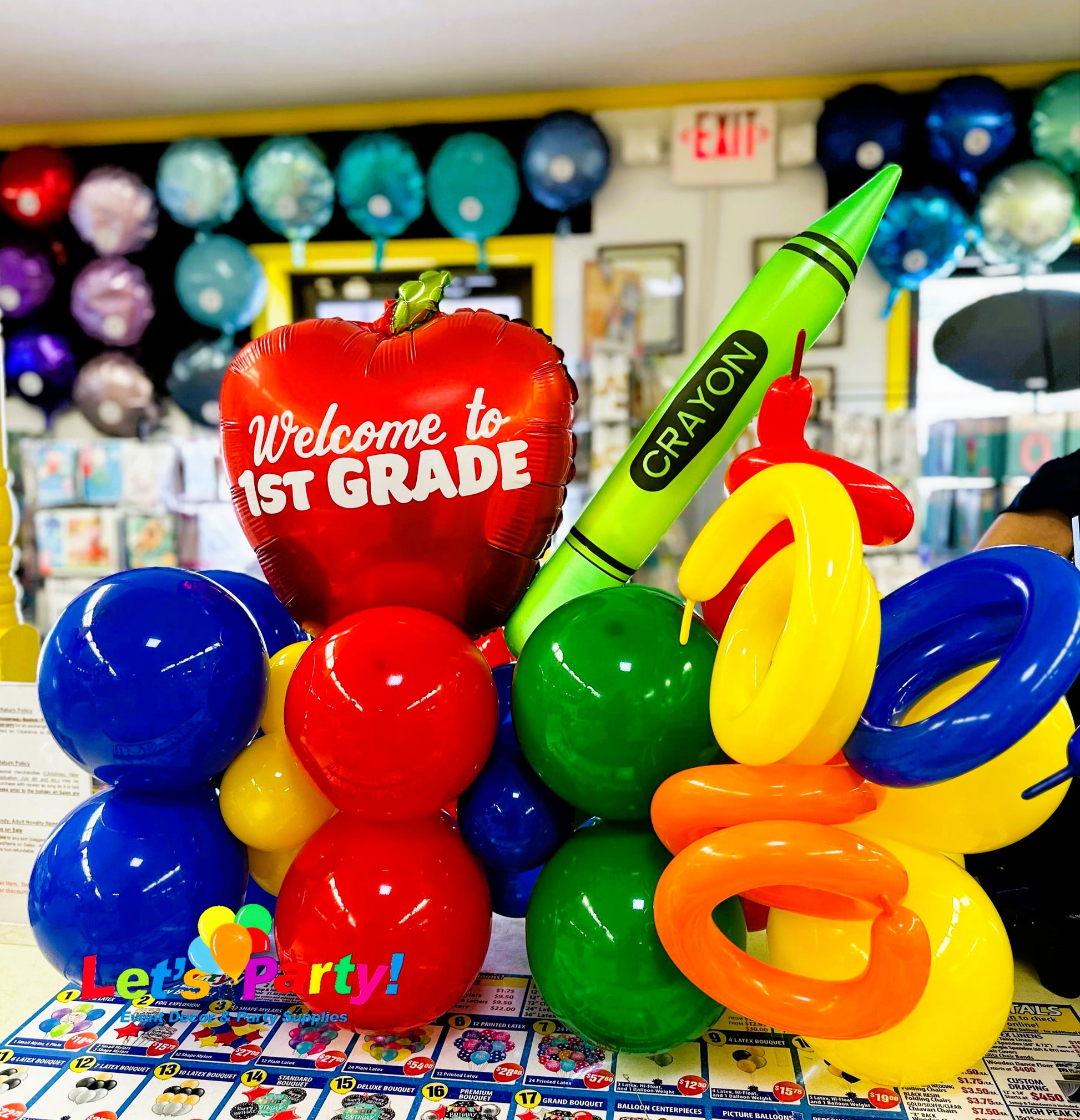 A colorful back to school balloon bouquet in a classroom setting, featuring a red apple-shaped balloon that says 'Welcome to 1st Grade', a large green crayon-shaped balloon, yellow balloons, blue and red balloons, and a small 'Let's Party!' sign at the base.